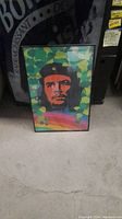 Framed colorful poster depicting Che Guevara leaning against a dark object on a concrete floor.