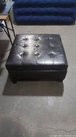 Large square black tufted storage ottoman with faux leather upholstery seen from the top and front.