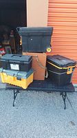 Photo of 5 miscellaneous empty tool boxes stacked on a black folding table outside a storage unit door.