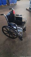 Side and angled views of manual wheelchair with black padded seat and back, chrome frame, pushed indoors on concrete floor.