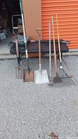 Seven yard tools displayed upright in front of an orange storage unit door showing variety and condition.