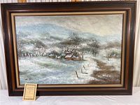 Framed oil painting showing snowy rural scene with houses and trees, signature visible on lower left corner.