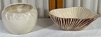 Two vintage pottery pieces side by side on a table with a neutral backdrop. Left is a cream-colored vase with leaf relief, right is a shell-shaped dish with brown and cream coloring.