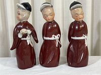 Front view of three ceramic religious monk figurines, each about 5.5 inches tall, with maroon robes and headwear, one holding a lamb.
