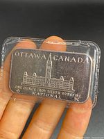 Front of the silver bar showing Ottawa Canada building engraving and purity markings in plastic case held by fingers.