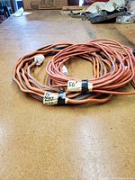 Two coiled orange heavy duty extension cords placed on a wooden surface, each about 50 feet long, with tape labeling length and gauge of one cord as 12 gauge.