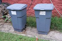 Front view of two gray plastic trash bins with lids and wheels placed outdoors on pavement next to grass and a red wall.