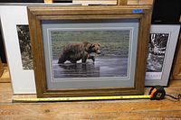 Photo capturing a framed color photograph of a bear by Peter Makuch in a wood frame with gray mats, measuring about 21 inches wide from tape measure at base.