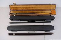 Three pool sticks laid out with their respective hard cases. One stick is in a brown velvet-lined wooden case, others in black plastic cases.