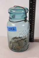 Ball brand blue-tinted glass jar filled with coins, placed next to a ruler for size reference.