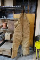 Full front view of the tan Carhartt bib overalls hanging, showing overall length and pockets.