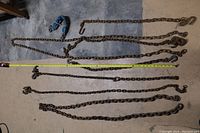 Set of seven heavy-duty metal chains laid out on concrete floor, showing varying lengths and hooks attached on ends