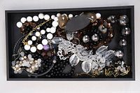 Photo showing an overview of costume jewelry pieces including bead necklaces and brooches in a black tray.