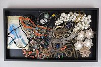 Top view showing a contained black tray full of assorted costume jewelry including pearl necklaces, bead necklaces, large decorative brooches with pearl centers, and mixed chains.