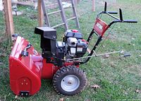 Left side view of snowblower showing red auger housing, black engine and handles, knobby tires