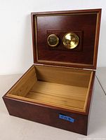 Open humidor box showing interior lined with lighter wood and brass hygrometer and humidifier attached to inside lid