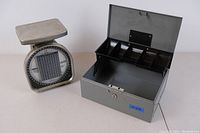 Mechanical postage scale and gray metal lock box opened to show compartments inside.