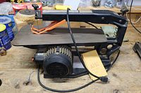 Black and orange electric scroll saw with a metal table surface and attached clamp. Power cord visible. Located on wooden workbench with tools and supplies in background.