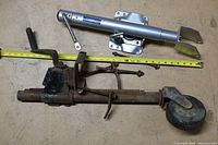 Photo showing two trailer jacks, one silver metallic with mounting base and crank, and one rusted with wheel and handle, placed on a floor with a yellow measuring tape beneath them for scale.