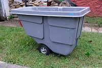Side view of a large gray plastic trash cart with two wheels on grass against a building background.
