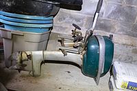 Side view of the entire vintage Mercury outboard motor showing green engine cover, mounting bracket and propeller on a shelf.