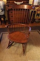 Full view of vintage wooden rocking chair showing spindle back, curved armrests, and traditional rockers on carpeted floor with wooden dresser background.