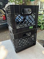 Two black plastic milk crates stacked vertically shown in outdoor daylight. Lattice sides and handles visible.