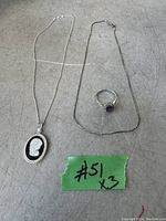 Two sterling silver chains laid flat, one with an oval black cameo pendant and one plain chain, plus a sterling silver ring with amethyst stone shown beside them. All rest on a textured surface.