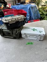 Three stacked storage boxes: black with distressed white lid on top, white metal box in the middle with green stenciling, and a green plastic box at the bottom with locking dial.