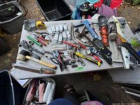 Overview of the hand tools including hammers, clamps, pliers, chisels, and assorted blades spread on a grey table.