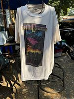 White Doobie Brothers T-shirt with colorful sunset and tree graphic, photographed hanging outdoors on a metal stand.