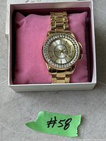Gold tone stainless steel wristwatch with crystal bezel resting on a pink cushion inside a white box with green lot number tag visible.