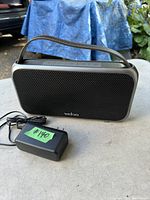 Veho retro Bluetooth speaker with black mesh front, brown leather strap and included charger seen outdoors with blue tarp background.