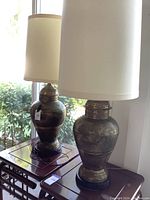 Two brass Moro jar lamps with off-white fabric cylindrical shades placed on wooden tables near a window.