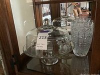 Photo showing clear glass cake stand with dome cover and Bohemian Czech crystal vase on a wood surface against cabinet background.