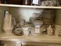 Full shelf view of stacked dinnerware including plates, cups, pitchers and bowls mostly wrapped in plastic.