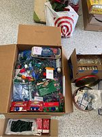 Wide view of two boxes; one with Christmas string lights, replacement bulbs, and egg carton with more bulbs or supplies; the other cardboard box contains a Christmas metal container