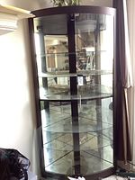 Tall corner curio cabinet with dark expresso finish, glass shelves and glass doors. The inner shelves and mirrored back visible. Overall cabinet intact but left glass door broken.
