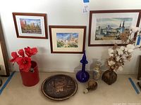 Wide shot of the lot showing three framed signed foreign cityscape pictures, Greek signed decorative plate, and five different vases including blue glass, red ceramic, and decorated beige vase.