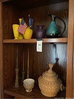 Overview of shelf with all items: teal glass pitcher with black handle; purple frosted glass vase; two yellow ceramic vases; carved wood candlesticks; white leaf-patterned cup; lidded wicker basket.