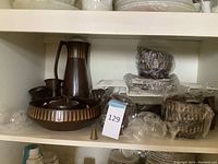 Wide shelf shot showing large brown stoneware pitcher, large bowl with geometric pattern, multiple smaller bowls and other dishes wrapped in plastic.