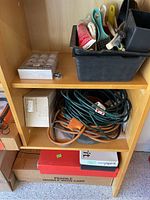 Garage shelf with plastic containers holding hand tools, jumping cables coiled on shelf below, and several boxes for hardware parts
