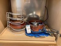 Photo showing stacked brown ceramic-coated pots and pans with glass lids and blue fabric protectors on a wooden shelf with a white metal rack visible to the left.