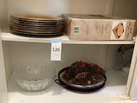 Shelf with 12 gold leaf finish plate charges stacked and a box, bottom shelf showing carved glass bowl and platters with pine cones on them.