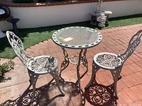 Full view of metal bistro set including round table with glass top and two chairs with rose design, showing outdoor tiled brick patio setting