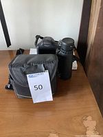 Photo shows Nikon camera body with attached zoom lens and accompanying black carrying case with yellow zipper on wooden surface.