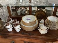 Stack of Noritake Manor Gold dinner plates, cups stacked alongside, with a sugar bowl visible.