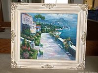 Full view of the framed landscape painting showing the coastal scene and ornate white and gold frame.