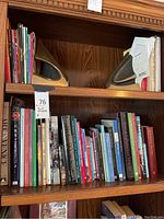 Wide view of two solid brass bookends placed on a wooden bookshelf along with various books on art, history, and culture.
