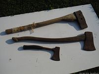 Top down view of two axes and one hatchet laid on a white surface outdoors showing wooden handles and rusted metal heads.